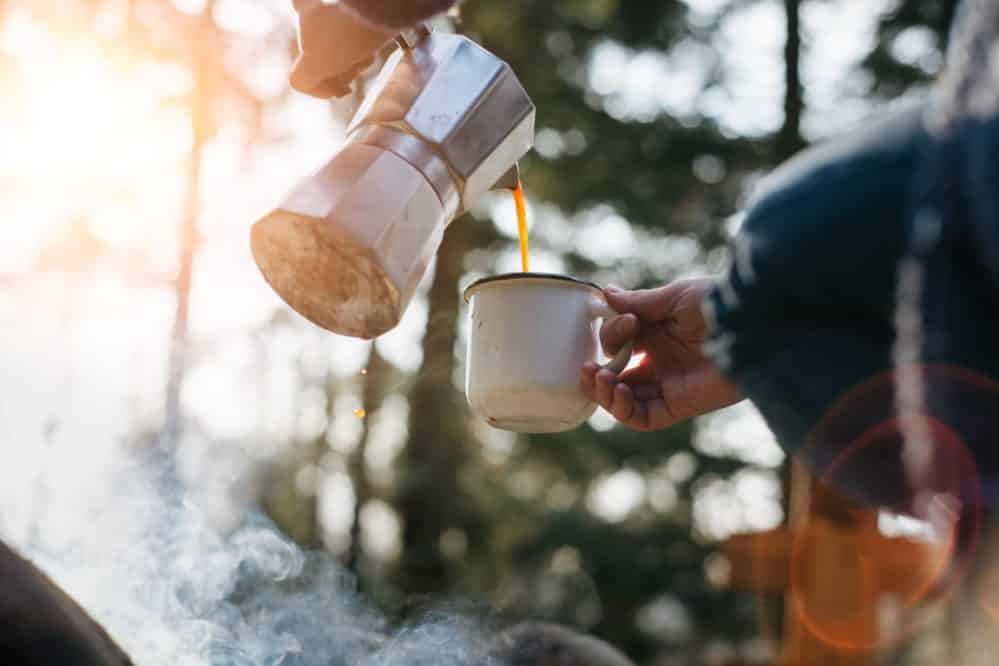 horizontal outdoors image of young woman pours itself hot beverage in mountains near to bonfire . girl sitting and holding a mug of coffee after hiking. travel and healthy lifestyle concept