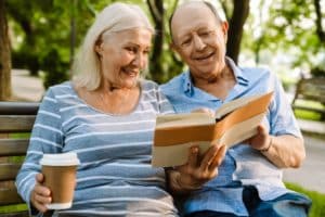 white senior couple drinking coffee and reading book while sitting on bench